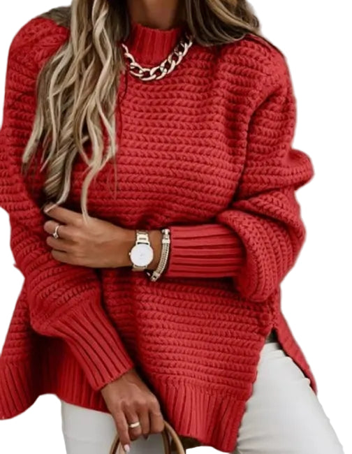 A woman wearing a chunky red knit sweater with a high mock neck and oversized sleeves, paired with white pants and gold jewelry, standing against a gray wall.