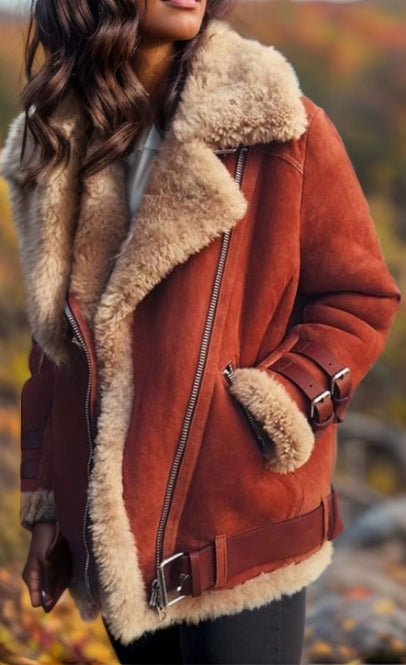 A woman wearing a brown suede aviator-style jacket with a thick beige shearling collar and lining, photographed outdoors against a blurred autumn background.