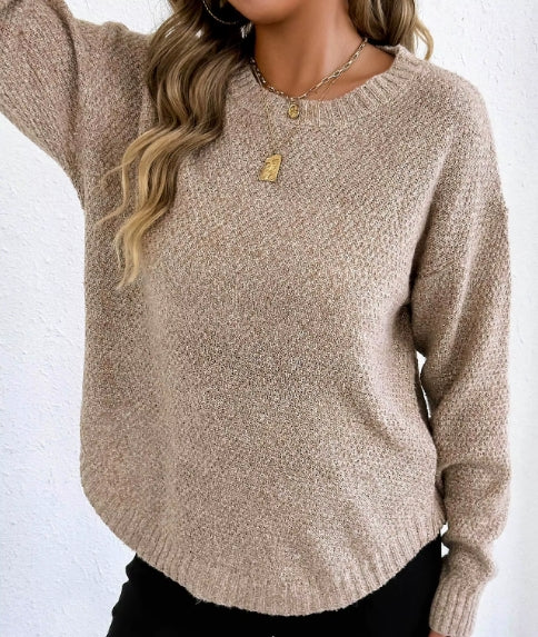 A woman wearing a beige textured knit crewneck sweater, paired with black pants and gold necklaces, against a white textured wall.