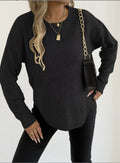 A woman wearing a dark charcoal gray textured knit sweater with black pants, accessorized with gold necklaces and a black shoulder bag with a gold chain strap.