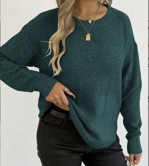 A woman wearing a dark teal green textured knit sweater with black pants and layered gold necklaces, shown from the chest down against a white wall.