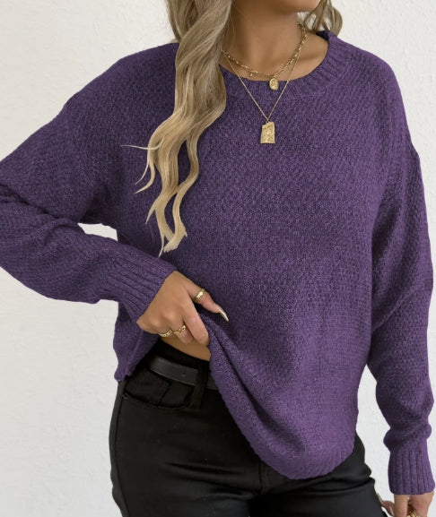 A woman wearing a textured purple knit sweater and black pants, shown from the neck down against a plain white wall.