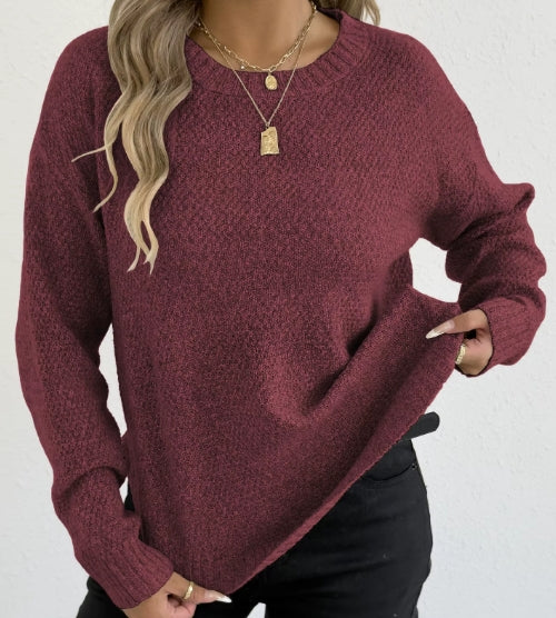 A woman wearing a textured, long-sleeve crewneck sweater in a heathered burgundy color, paired with black pants and layered gold necklaces, set against a plain white wall.