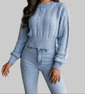 A woman wearing a light blue cable-knit sweater and matching jeans, holding a brown faux-leather clutch bag with fringe and silver-tone hardware.