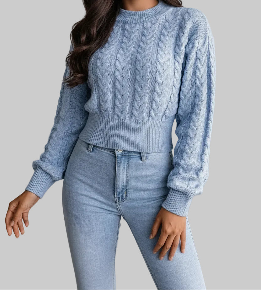 A woman wearing a light blue cable-knit sweater and matching jeans, holding a brown faux-leather clutch bag with fringe and silver-tone hardware.