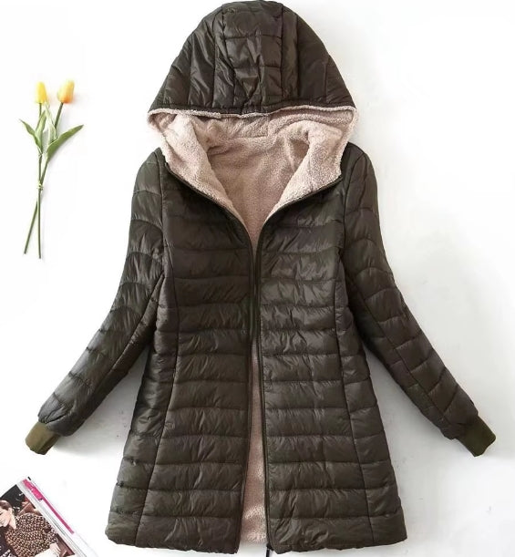 A women's quilted hooded puffer jacket in dark olive green with a soft beige fleece lining, displayed flat against a white background with decorative tulips and a magazine.
